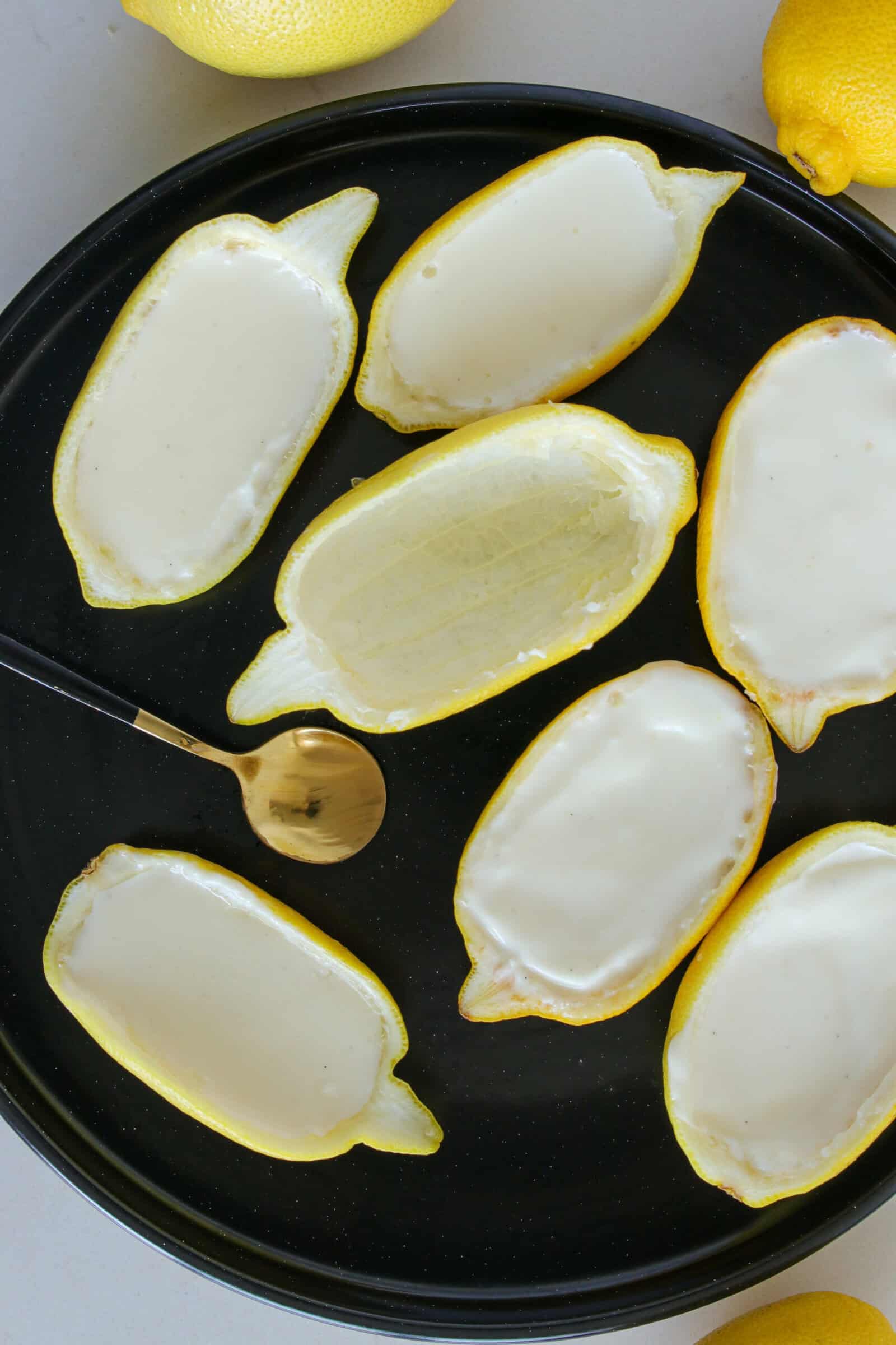 Halved lemons with their insides scooped out are filled with a creamy posset, arranged on a black plate with a gold spoon nearby. Whole lemons are partially visible at the edge of the plate.