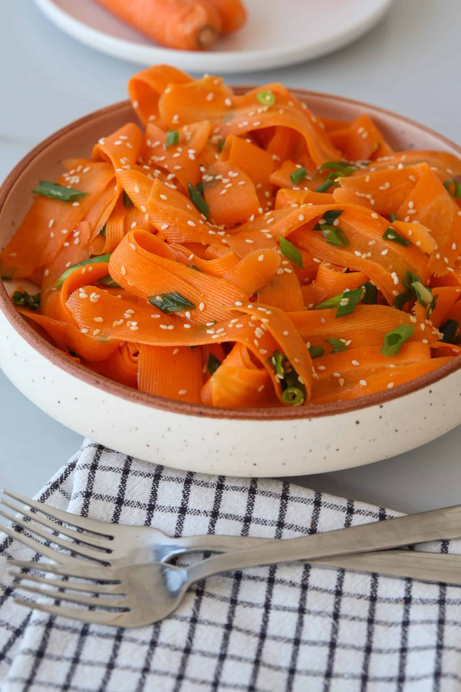 A bowl of carrot ribbon salad garnished with chopped herbs and sesame seeds, placed on a checkered napkin with two forks beside it; a carrot is on a plate in the background.