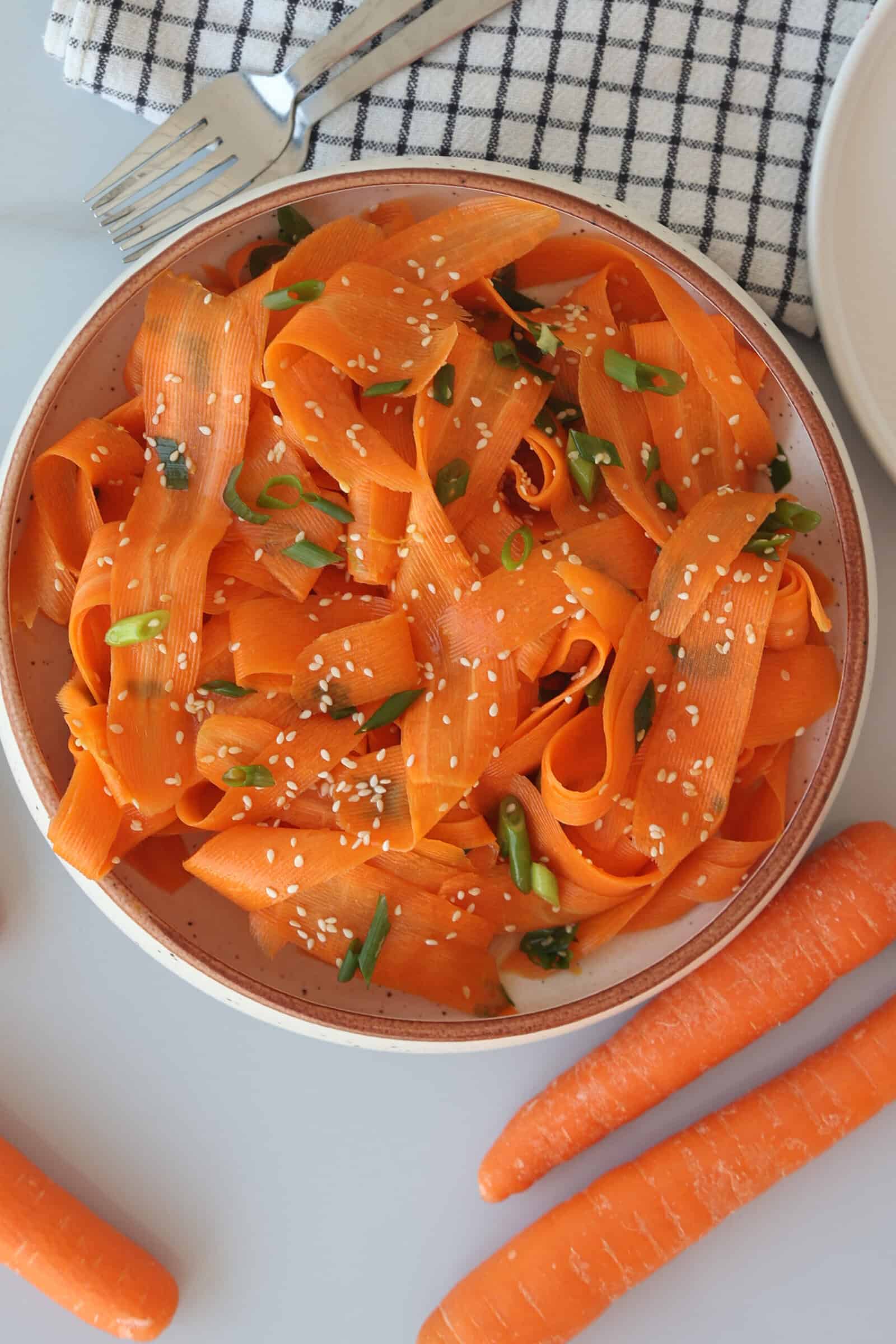A bowl of carrot ribbons garnished with sesame seeds and chopped green onions sits on a table beside whole carrots, a fork, and a checkered cloth napkin.
