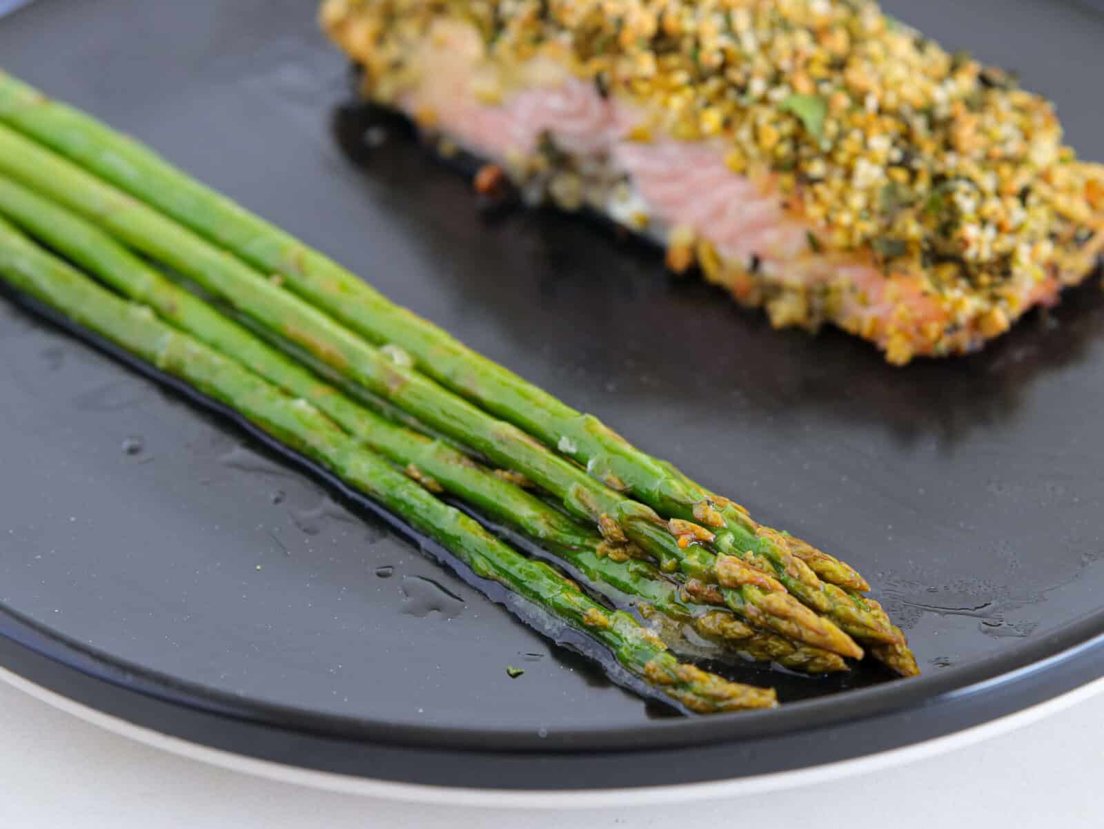 A close-up of cooked asparagus spears and a piece of herb-crusted salmon on a black plate. The asparagus is arranged neatly in the foreground.