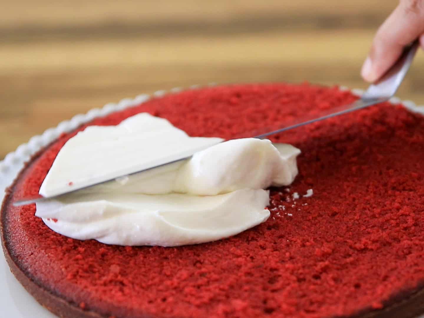 A hand uses an offset spatula to spread white cream frosting over a single layer of red velvet cake on a plate, with a blurred wooden background.