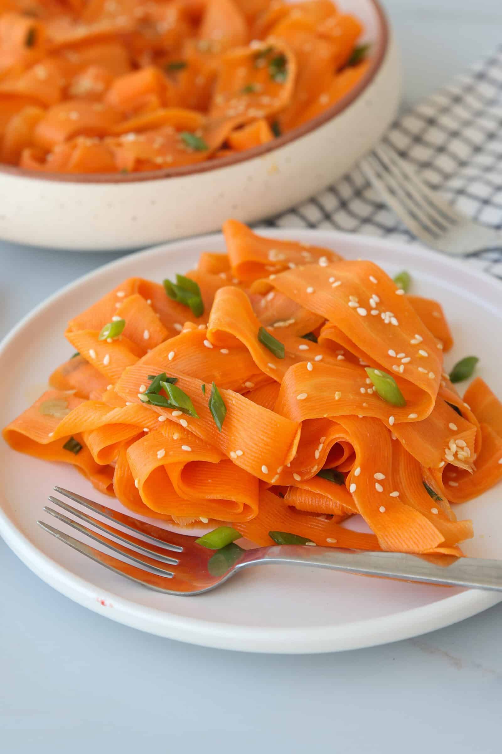 A white plate with wide carrot ribbons garnished with sesame seeds and chopped green onions, with a fork on the side. In the background, more carrot ribbons are visible in a shallow dish.