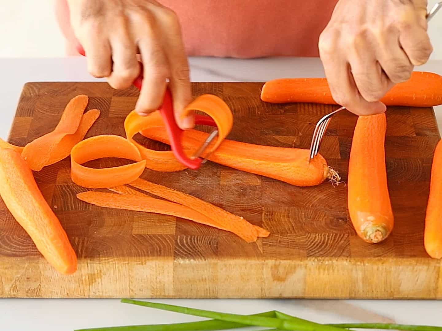 A person uses a vegetable peeler to peel a carrot on a wooden cutting board, with peeled carrot skins and whole carrots scattered around.