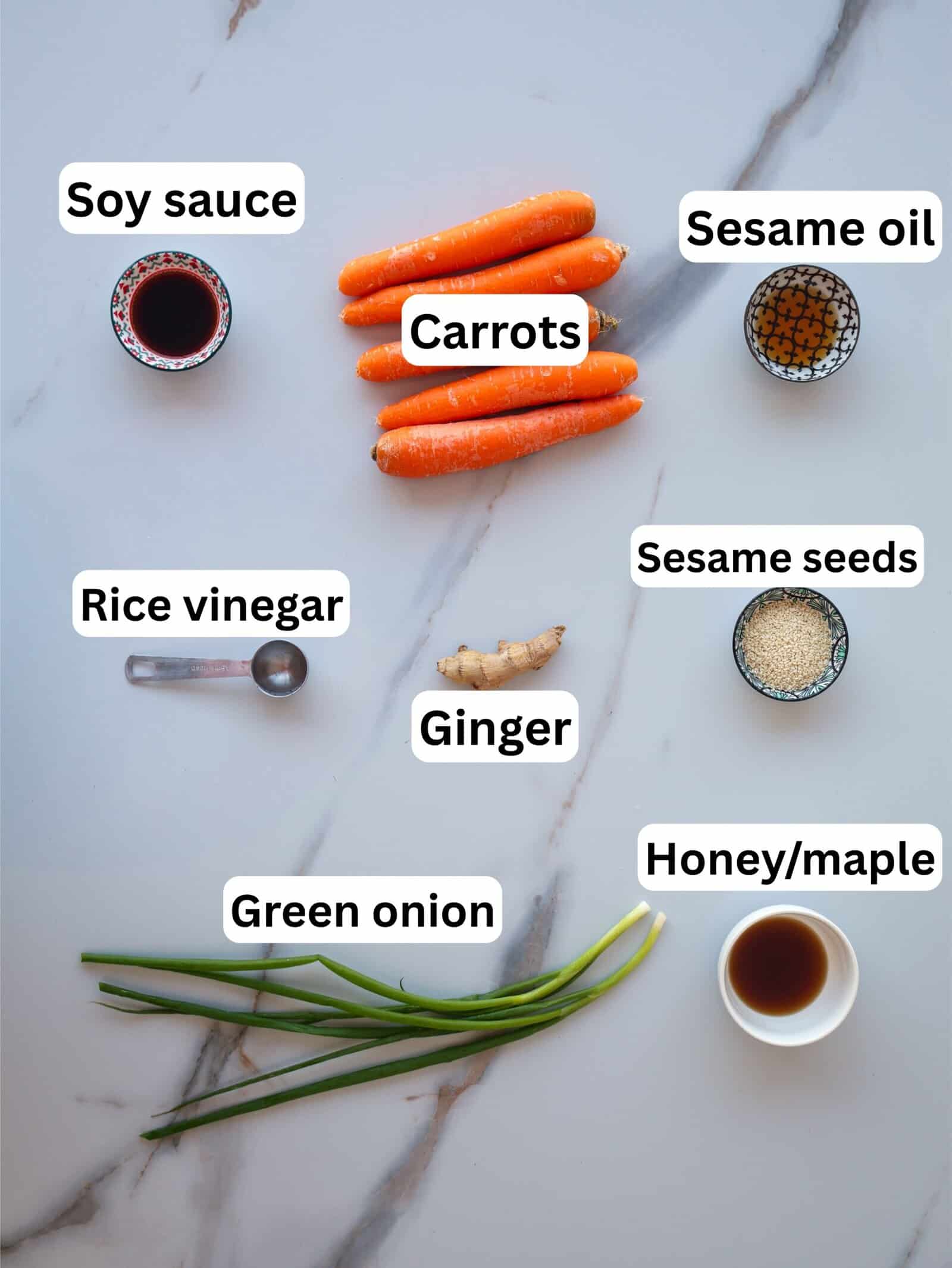 Ingredients for a carrot salad arranged on a marble surface, including labeled bowls of soy sauce, sesame oil, rice vinegar, sesame seeds, honey/maple syrup, and fresh carrots, ginger, and green onion.