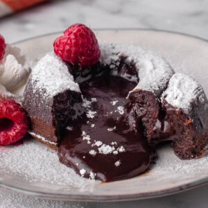 A chocolate lava cake with a gooey, molten center flows onto a plate. The cake is dusted with powdered sugar and garnished with fresh raspberries and a dollop of whipped cream.