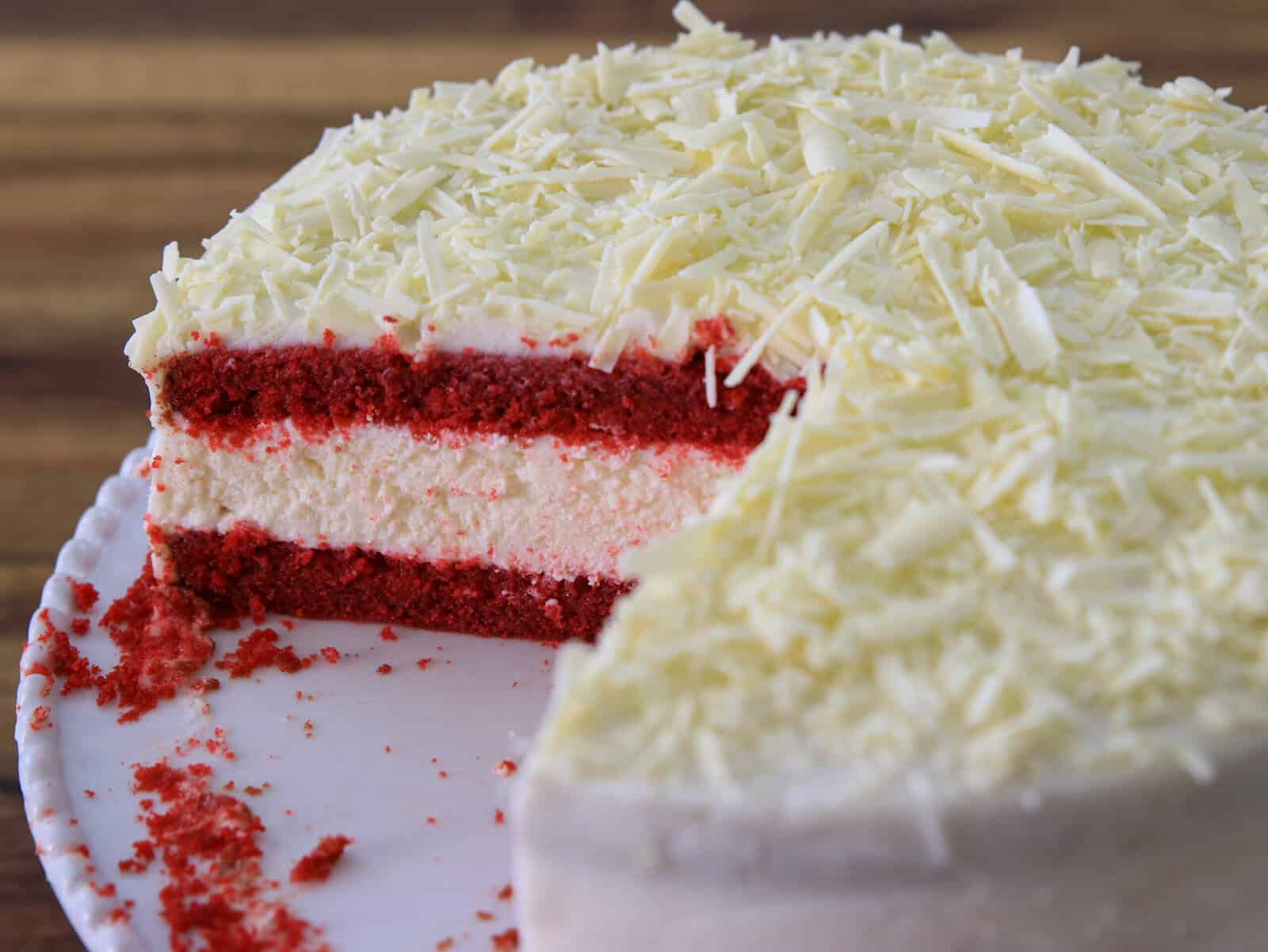 A partially sliced red velvet cake with white frosting and white chocolate shavings on top, revealing two layers of red cake and a creamy white filling inside.