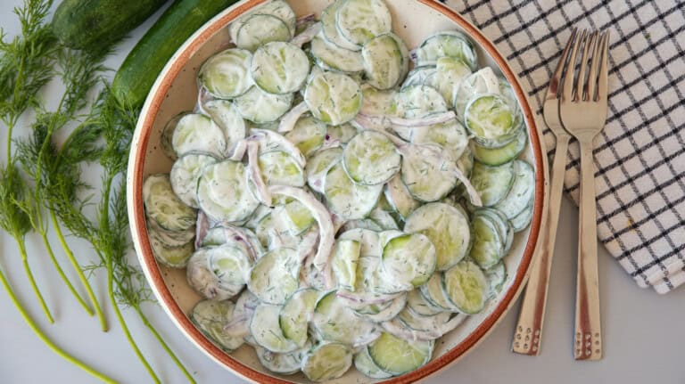 A bowl of creamy cucumber salad with sliced cucumbers and red onions, topped with fresh dill. The bowl is next to a fork and knife on a checkered napkin, with dill sprigs on the side.