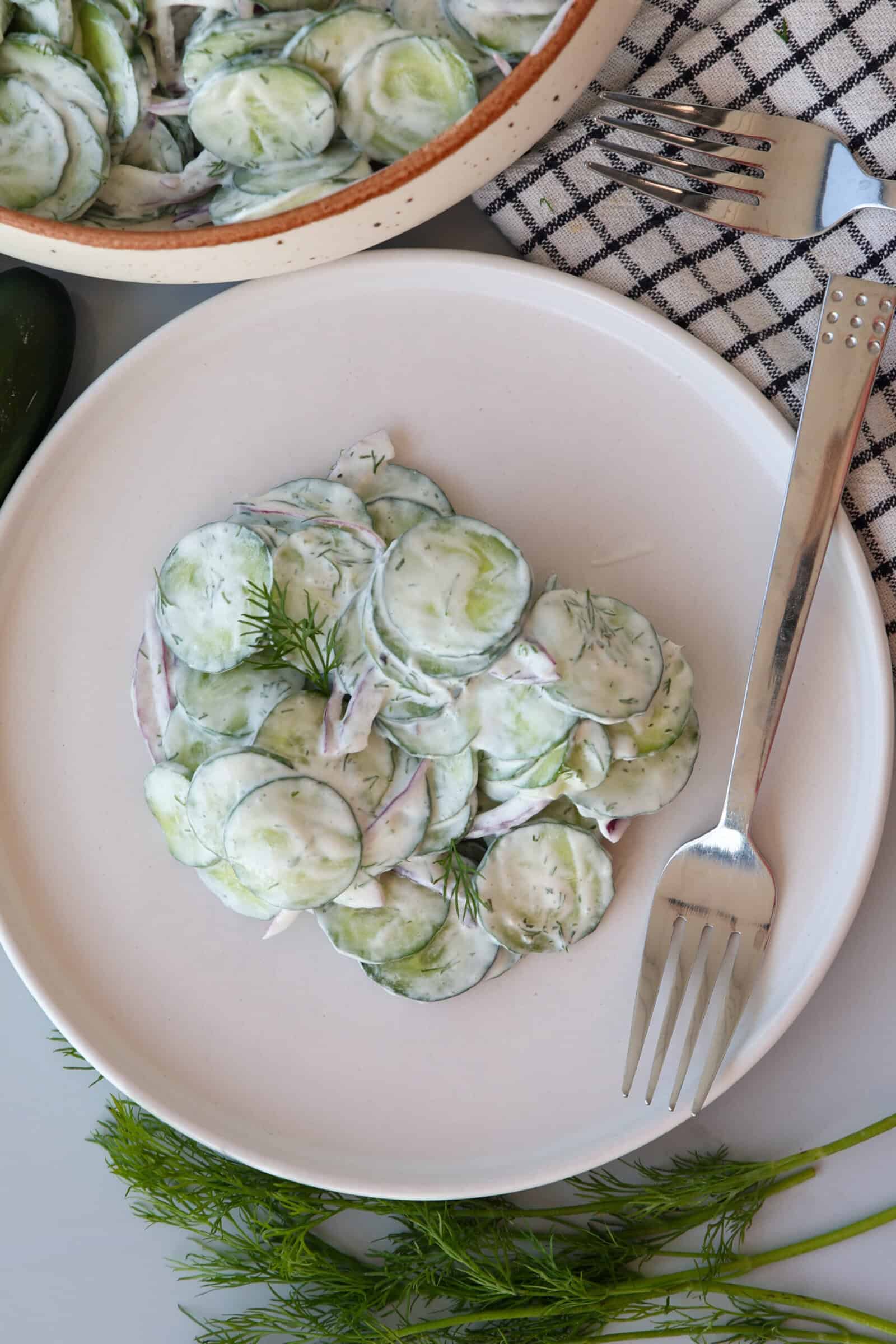 A plate of creamy cucumber salad garnished with dill, served with a fork. The salad includes thinly sliced cucumbers and onions in a creamy dressing. Fresh dill and a checkered napkin are nearby.