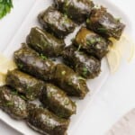 A plate of stuffed grape leaves garnished with chopped parsley, served with lemon wedges on a white rectangular dish.