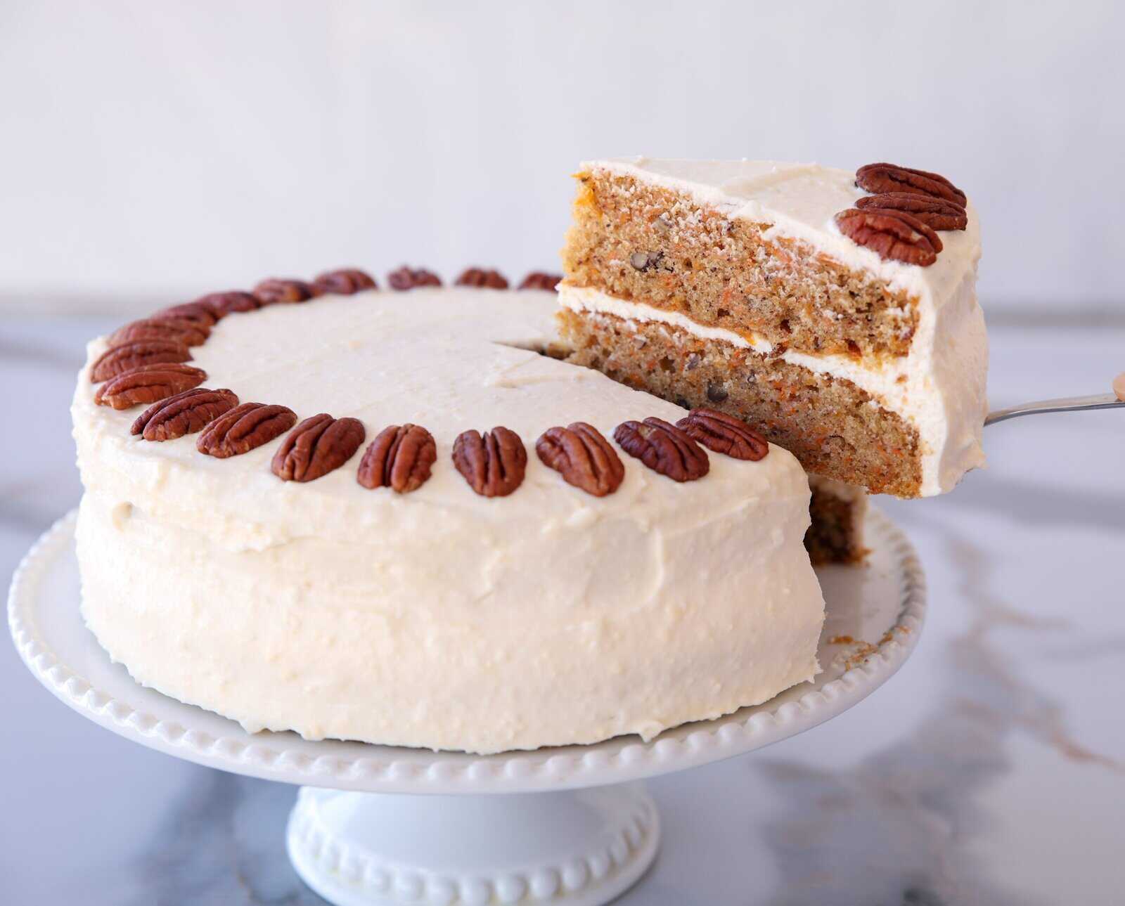 A round carrot cake with white cream cheese frosting, topped with whole pecans around the edge. A slice is being lifted from the cake, revealing the moist, nut-filled interior. The cake sits on a white cake stand.