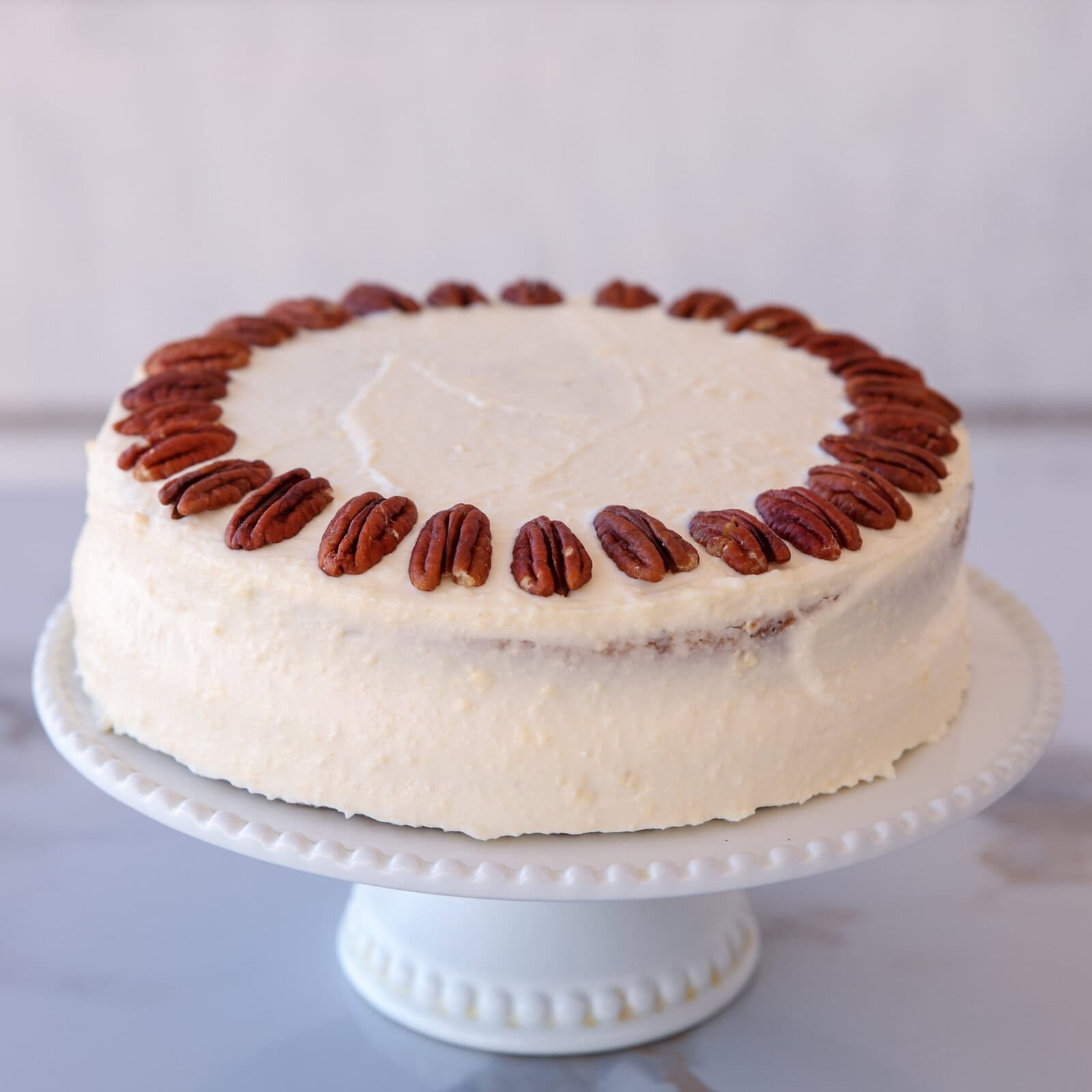 A round cake with white frosting, decorated with a ring of whole pecans around the top edge, sits on a white pedestal cake stand against a light background.