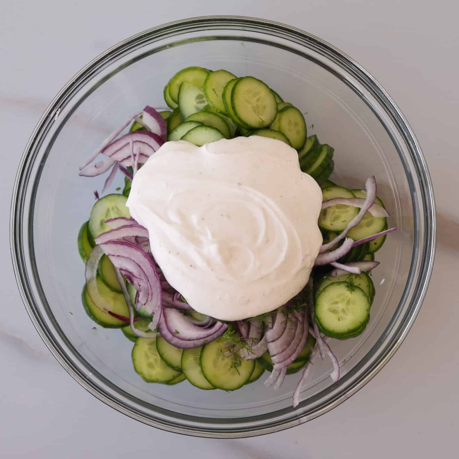 A glass bowl filled with sliced cucumbers and red onions, topped with a generous dollop of creamy white dressing, sits on a light-colored surface.