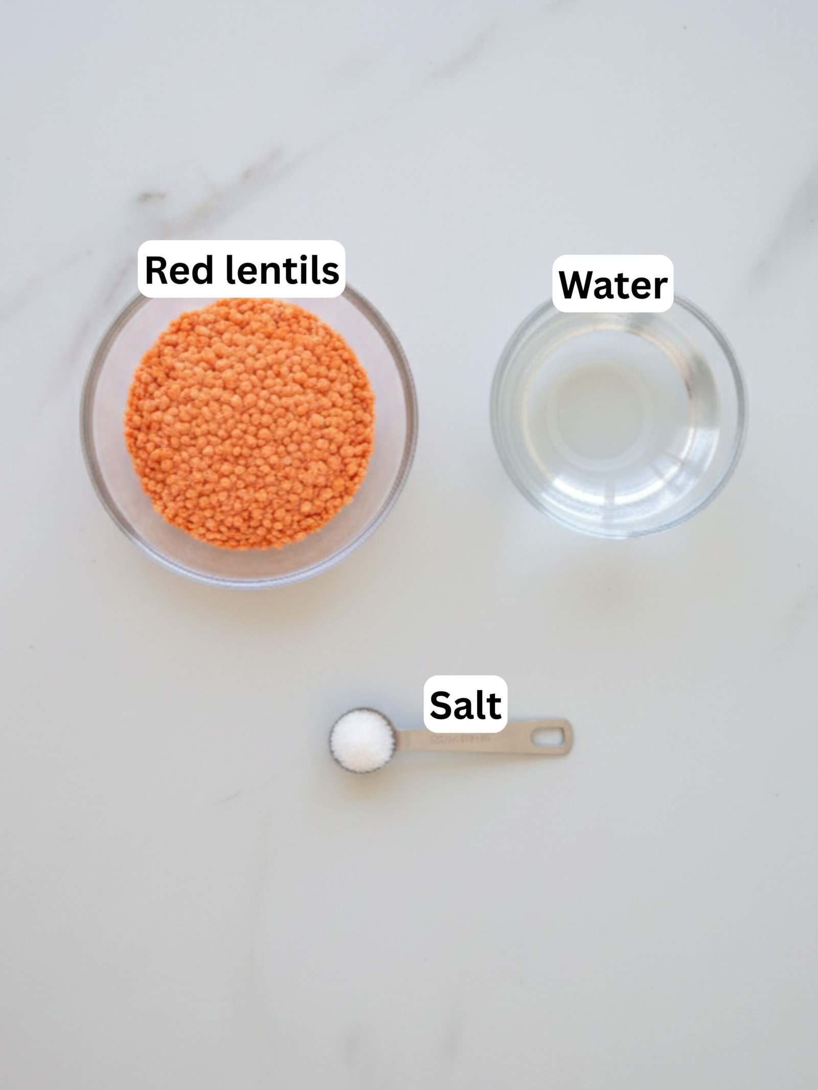 A bowl of red lentils, a bowl of water, and a measuring spoon with salt are arranged on a white surface. Each item is labeled: "Red lentils," "Water," and "Salt.