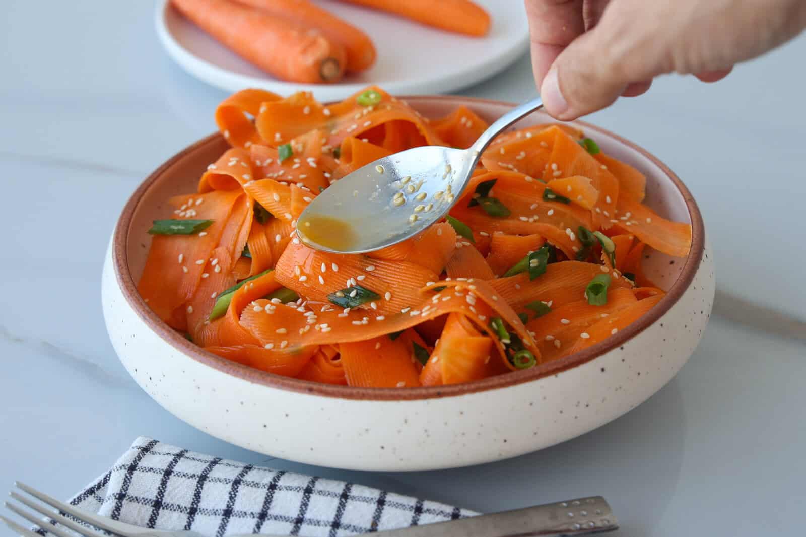 A hand drizzles dressing with a spoon over a bowl of ribboned carrot salad, garnished with sesame seeds and green onions. A plate with whole carrots is in the background, next to a fork and napkin.