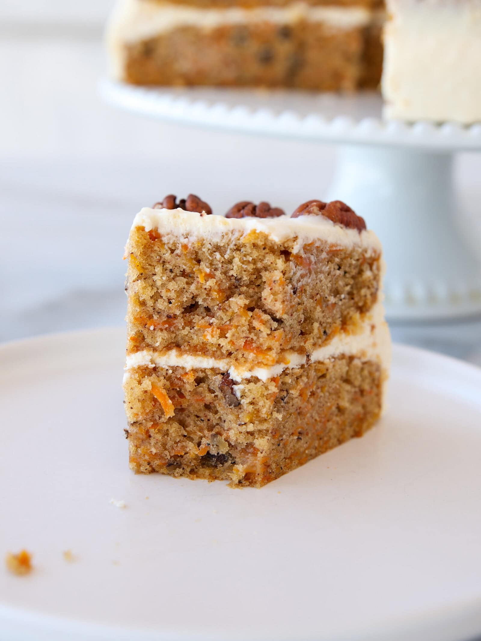 A close-up of a slice of carrot cake with cream cheese frosting, topped with pecans, sitting on a white plate. The cake is moist with visible pieces of carrot and nuts.