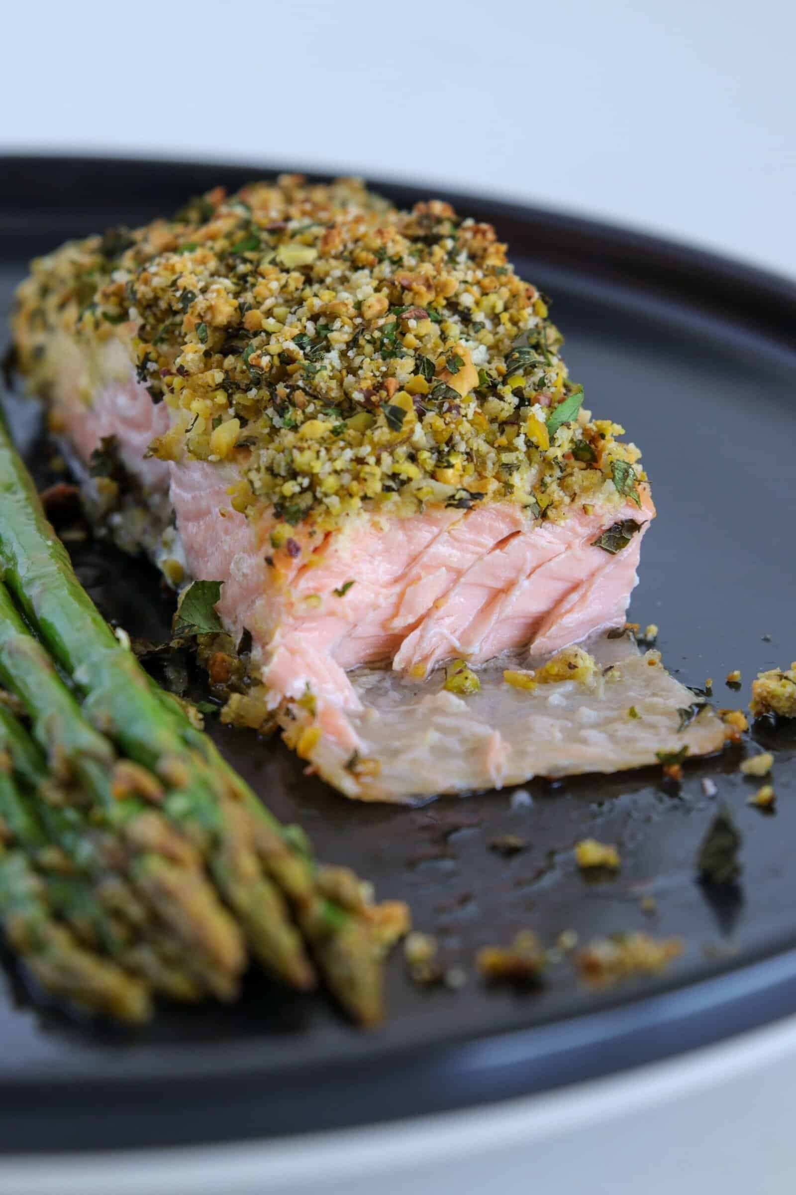 A close-up of a baked salmon fillet with a golden herb, pistachios and breadcrumb crust, partially flaked to reveal the tender pink inside, served with roasted asparagus spears on a dark plate.