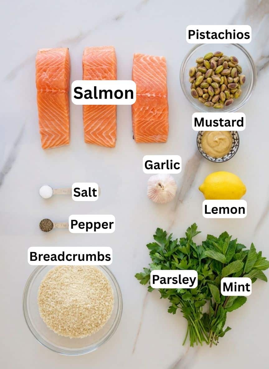 Three raw salmon fillets with bowls of pistachios, mustard, breadcrumbs, salt, pepper, a head of garlic, half a lemon, and fresh parsley and mint sprigs arranged on a white marble surface.