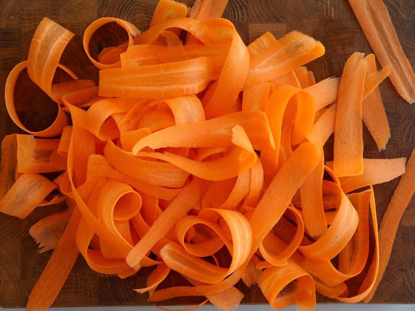 A pile of freshly peeled carrot ribbons lies on a wooden cutting board, showcasing vibrant orange colors and various curled and flat strips.