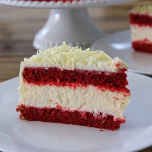 A slice of red velvet cheesecake cake with creamy white frosting and white chocolate shavings on top, served on a white plate. Another slice and a cake stand are visible in the background.