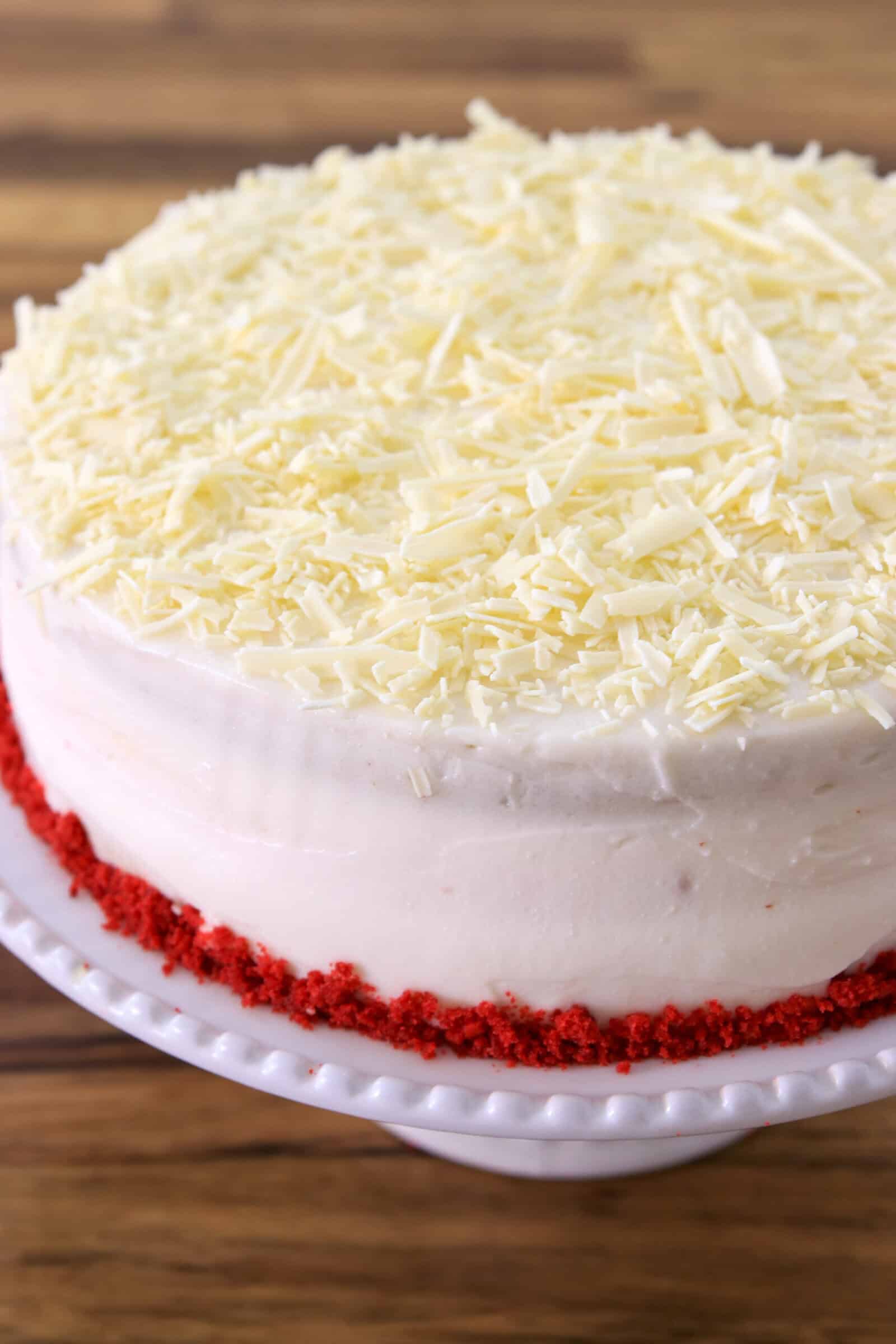 A round cake with white frosting, topped with white chocolate shavings and a border of red cake crumbs around the base, displayed on a white cake stand.