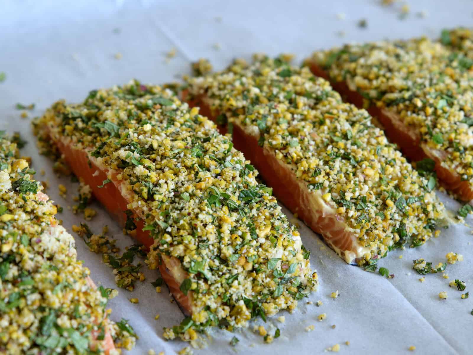 Four raw salmon fillets are lined up on parchment paper, topped with a crumbly herb and pistachio breadcrumb crust, ready for baking.