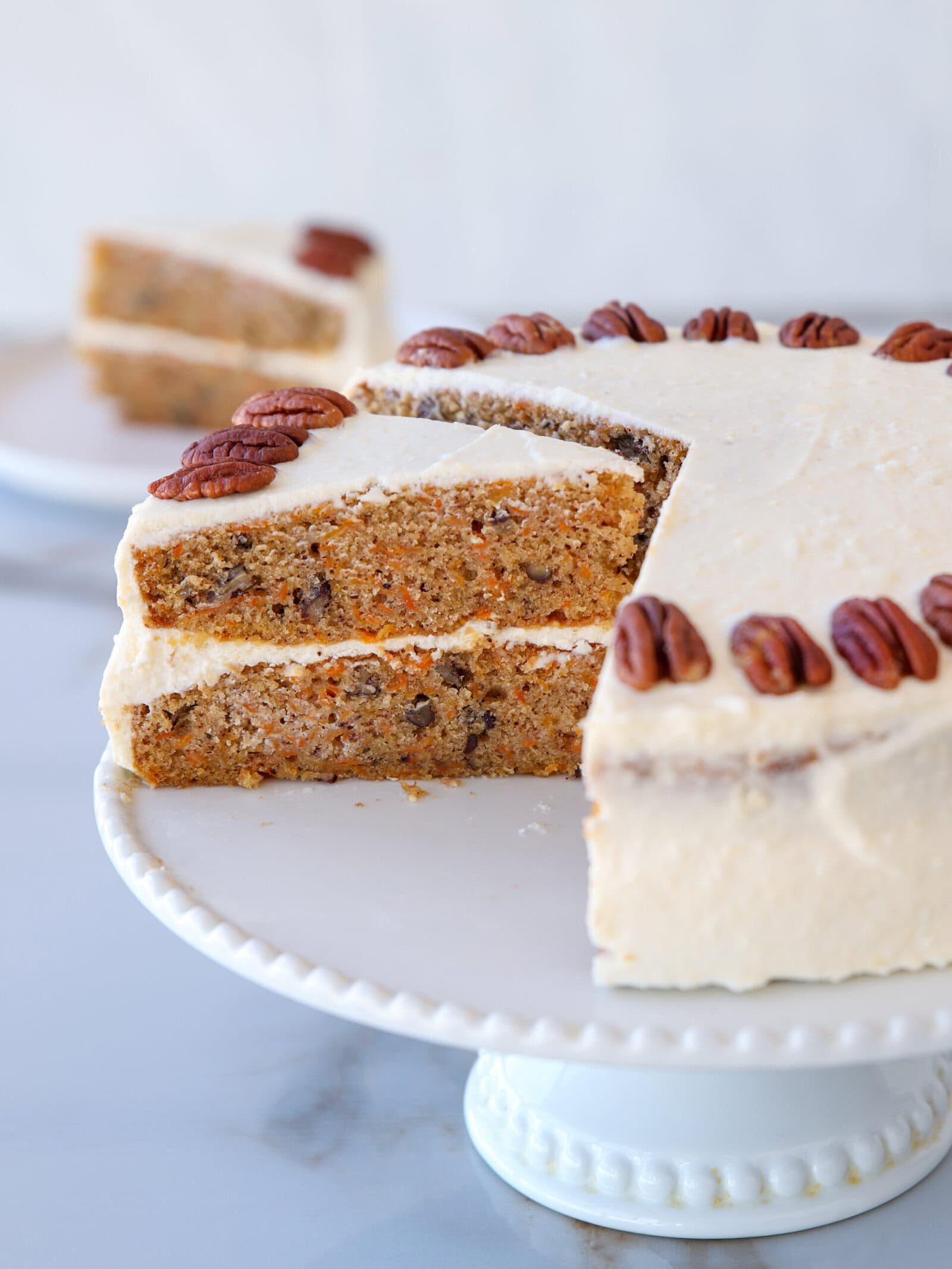 A sliced carrot cake with cream cheese frosting and pecans on top sits on a white cake stand. One slice is slightly pulled out, showing moist cake layers with nuts and carrot pieces inside.