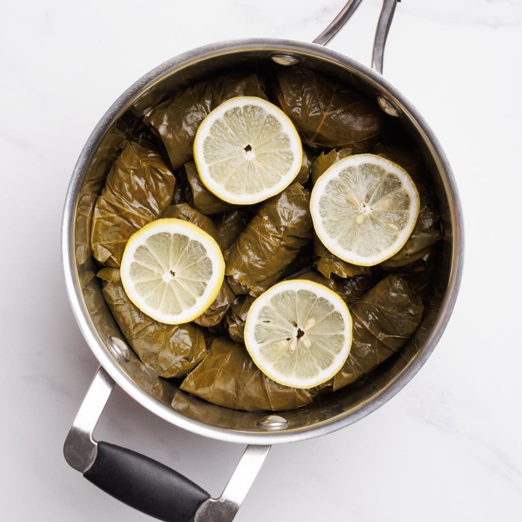 A pot filled with stuffed grape leaves, neatly arranged and topped with four slices of fresh lemon on a white surface.