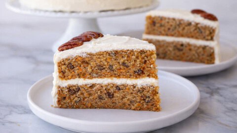 A slice of carrot cake with cream cheese frosting and a pecan on top, set on a white plate, with another slice and a whole cake in the background.