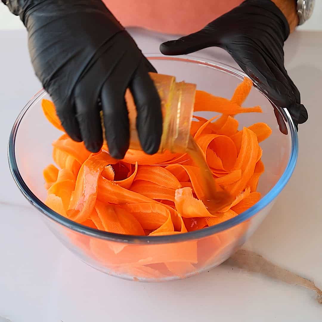 A person wearing black gloves pours liquid dressing from a jar onto a bowl of thinly sliced carrot ribbons on a white countertop.