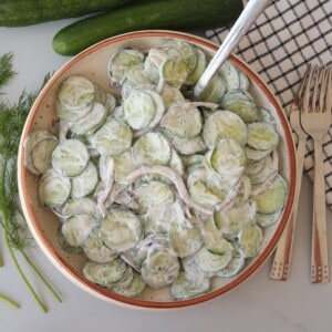 A bowl of creamy cucumber salad with sliced cucumbers, red onions, and dill in a creamy dressing. A spoon is in the bowl, with fresh dill, cucumbers, a napkin, and forks nearby.