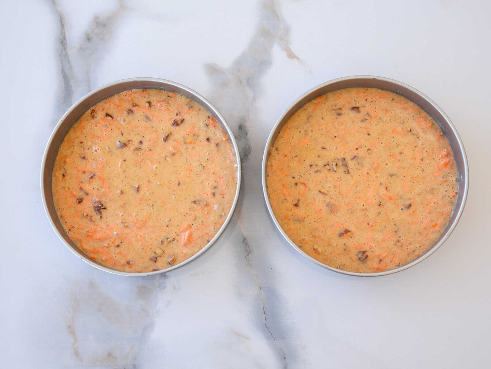 Two round cake pans filled with unbaked carrot cake batter containing visible shredded carrots and nuts, placed on a white marble surface.