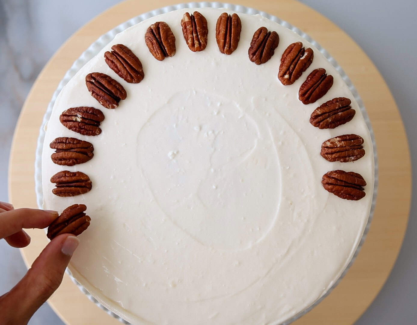 A hand places a pecan on a white frosted cake, which is decorated with a ring of pecans along the edge, leaving part of the circle unfinished. The cake sits on a light wooden surface.