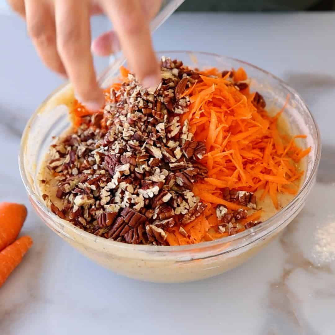 A hand reaches into a glass bowl filled with cake batter, shredded carrots, and chopped pecans, ready to be mixed. Two whole carrots lie on the marble countertop beside the bowl.