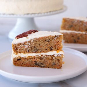A slice of carrot cake with cream cheese frosting and a pecan on top, served on a white plate. A whole frosted cake and another slice are visible in the background.