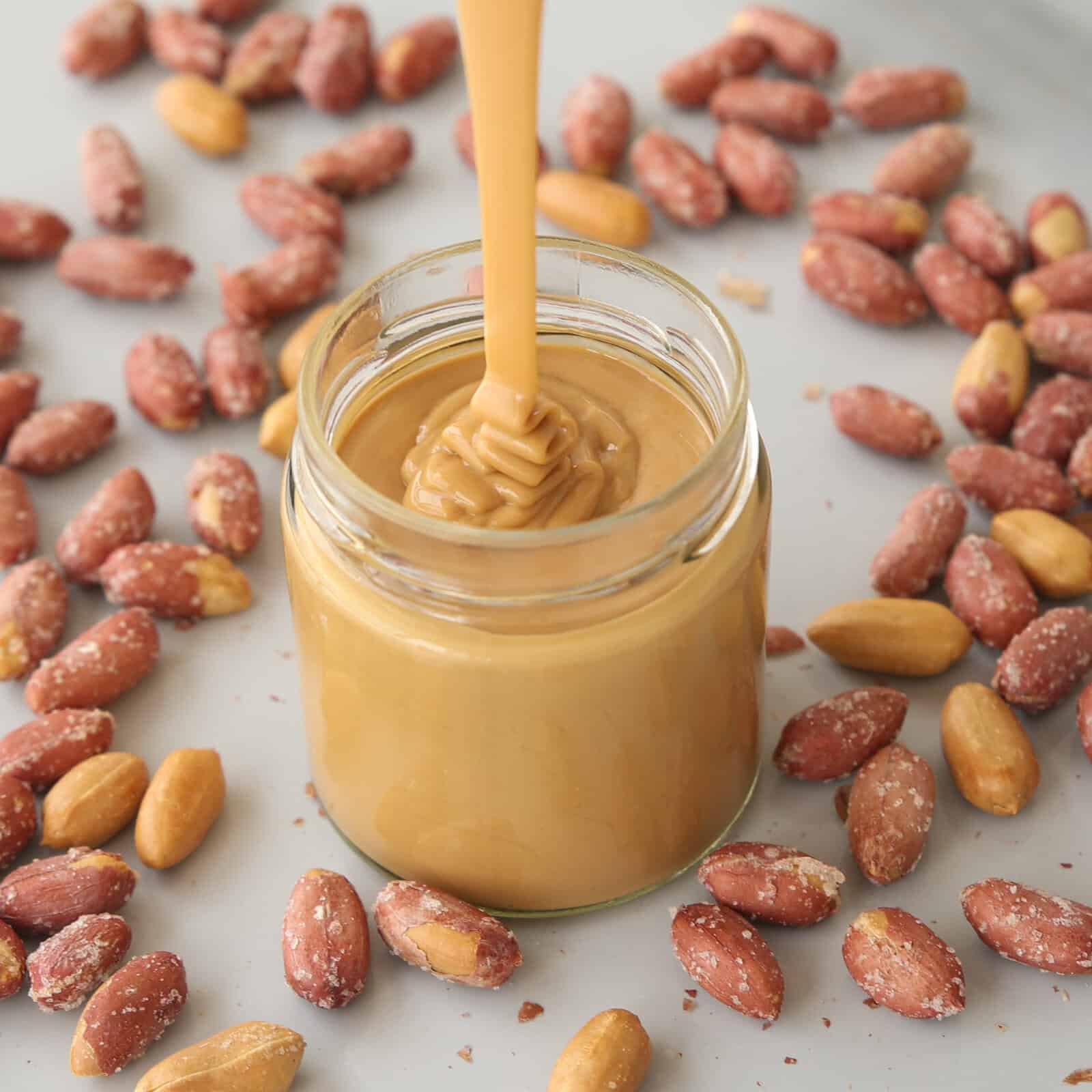 A stream of creamy peanut butter is being poured into a glass jar surrounded by salted peanuts on a light surface.
