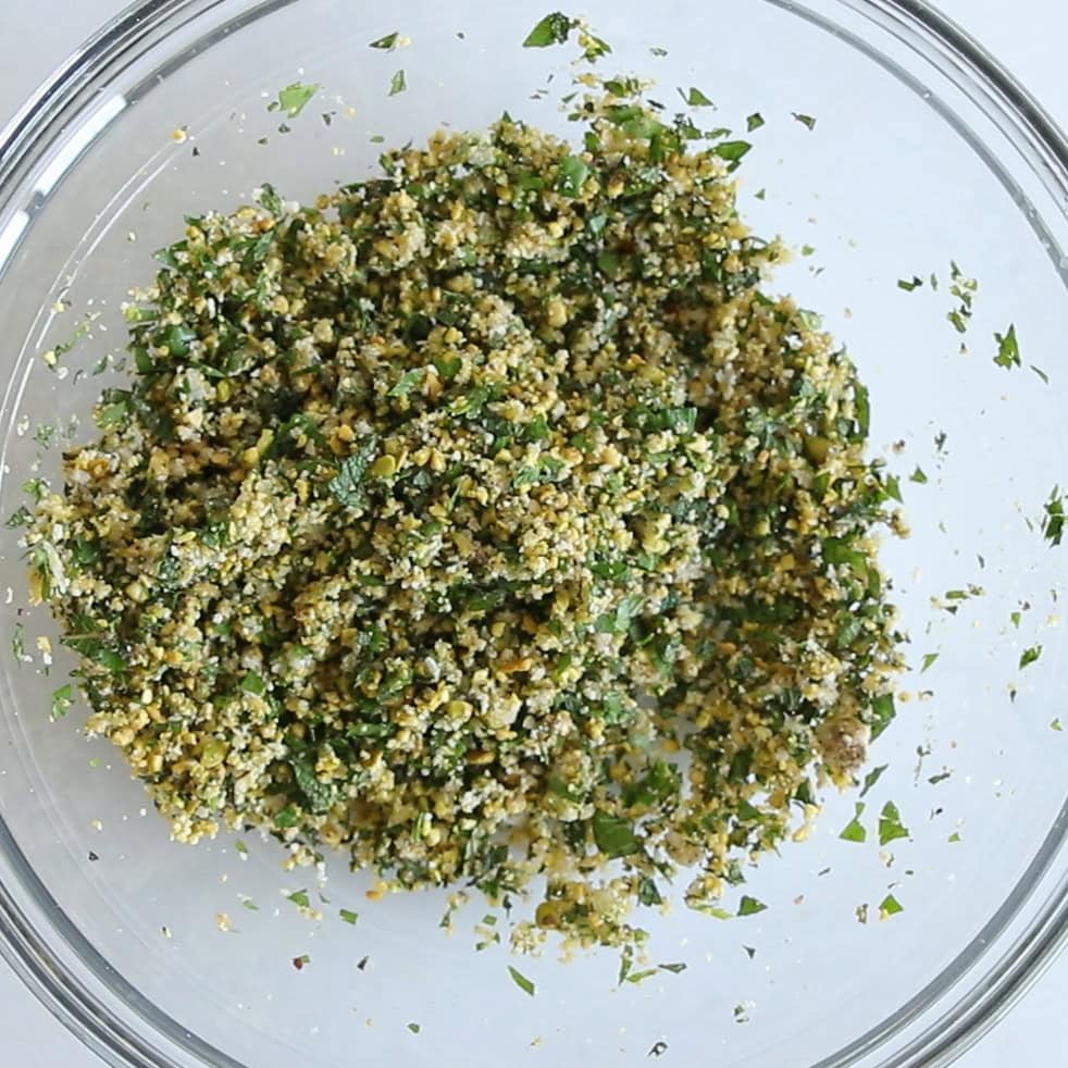 A clear glass bowl containing a mixture of finely chopped green herbs and breadcrumbs, evenly blended, on a white surface.