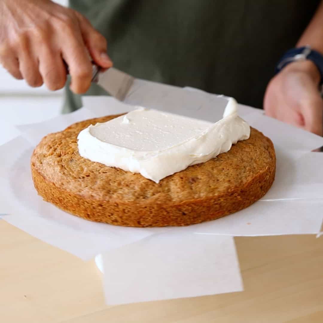 A person spreads white frosting on a round, unfrosted cake using an offset spatula. The cake sits on parchment paper atop a wooden surface.