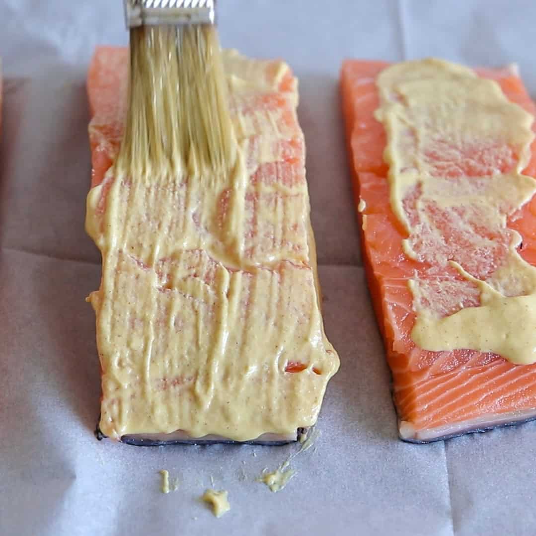 A close-up of a brush spreading a creamy mustard sauce over a raw salmon fillet on a sheet of parchment paper, with another salmon fillet beside it.
