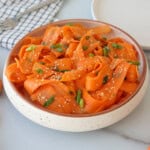 A bowl of carrot ribbons garnished with sliced green onions and sesame seeds sits on a white countertop, with a fork and a checkered napkin in the background.