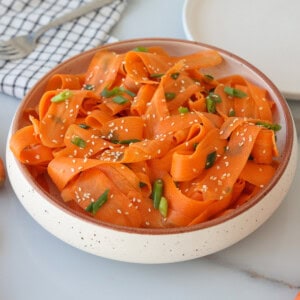 A bowl of carrot ribbons garnished with sliced green onions and sesame seeds sits on a white countertop, with a fork and a checkered napkin in the background.