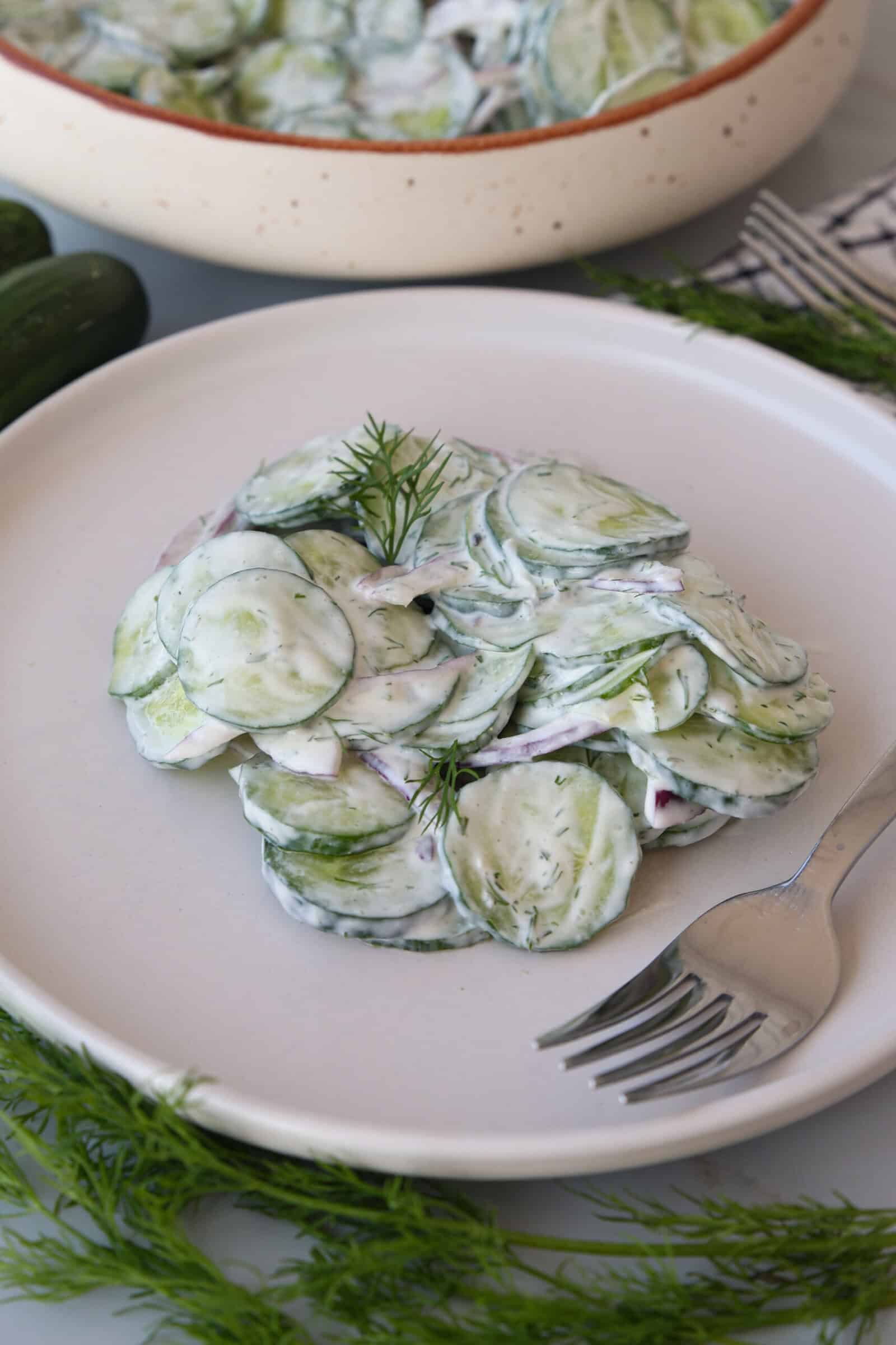 A plate of creamy cucumber salad garnished with dill, served on a white plate with a fork beside it. Fresh dill and part of a serving bowl are visible in the background.