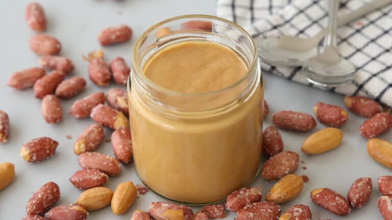 A glass jar filled with creamy peanut butter is surrounded by salted peanuts on a light surface, with a checkered cloth and two metal spoons in the background.