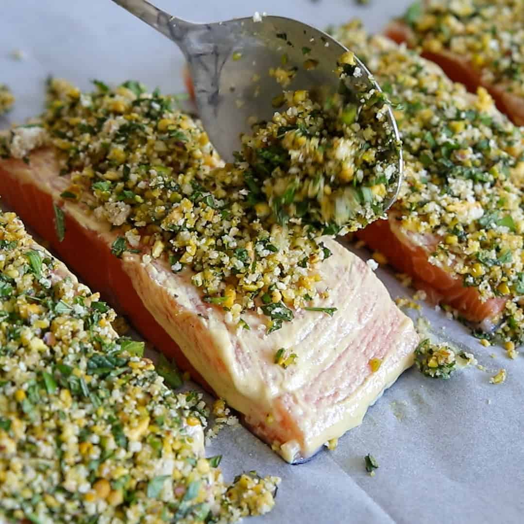 A spoon sprinkles a breadcrumb, herb, and nut mixture onto raw salmon fillets lined on parchment paper, preparing the fish for baking.