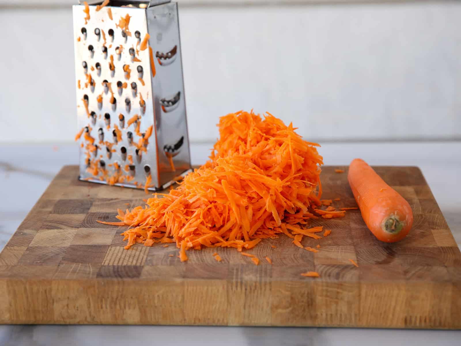 A pile of freshly grated carrots sits on a wooden cutting board next to a whole carrot and a metal box grater.