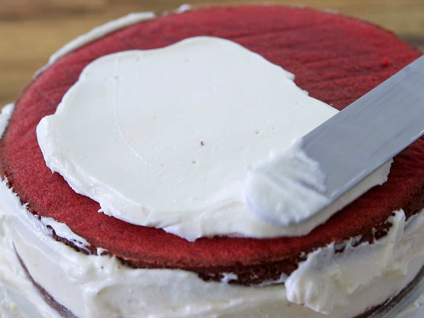 A spatula spreads a frosting over a red velvet cheesecake cake layers, with more frosting already applied to the cake’s sides and bottom layers.