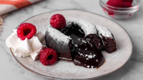A chocolate lava cake with liquid chocolate center oozing out, topped with powdered sugar, served with whipped cream and fresh raspberries on a white plate. A small bowl of raspberries is in the background.
