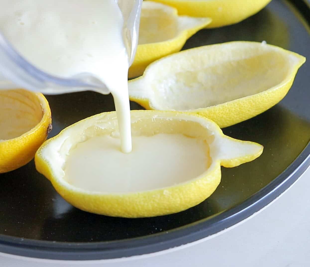 Creamy white liquid is being poured from a container into a hollowed-out lemon half, which is placed on a black plate with other empty lemon halves.
