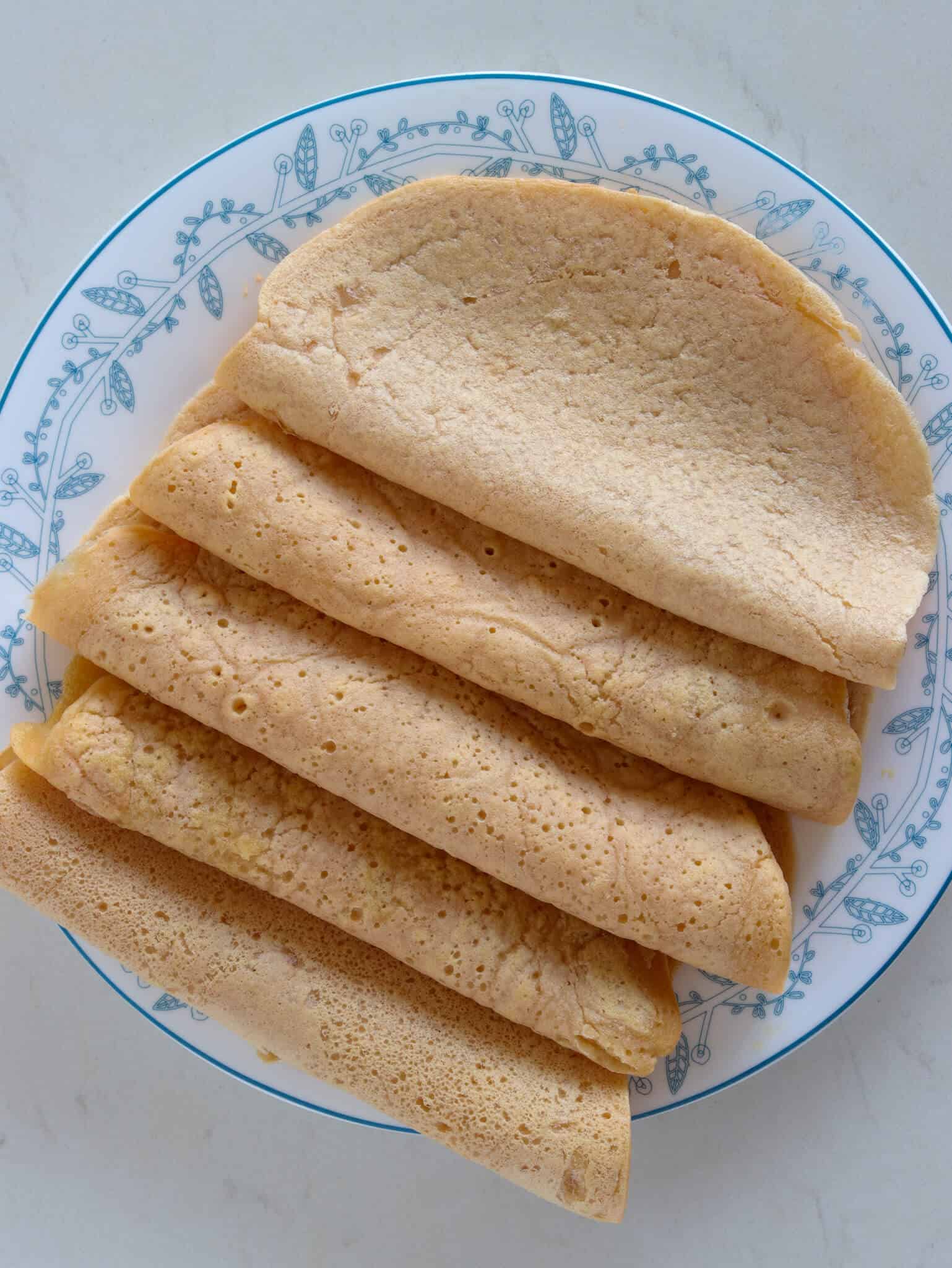 A plate with five folded, thin, golden-brown lentil wraps arranged in a fanned-out pattern on a white plate with a blue floral border.
