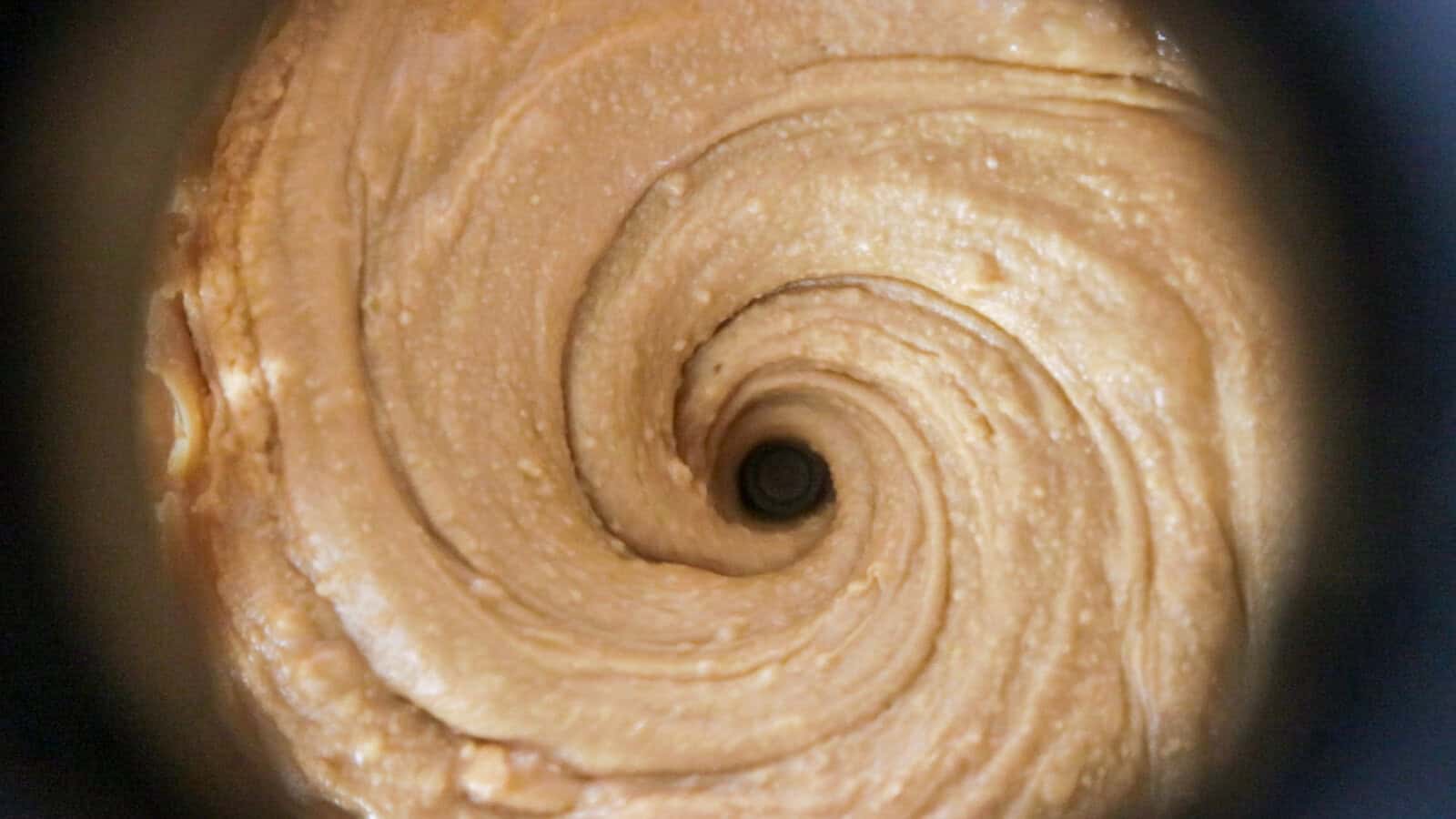 Close-up view of creamy peanut butter being swirled in a circular motion, creating a spiral pattern with a central vortex. The texture appears smooth and thick.
