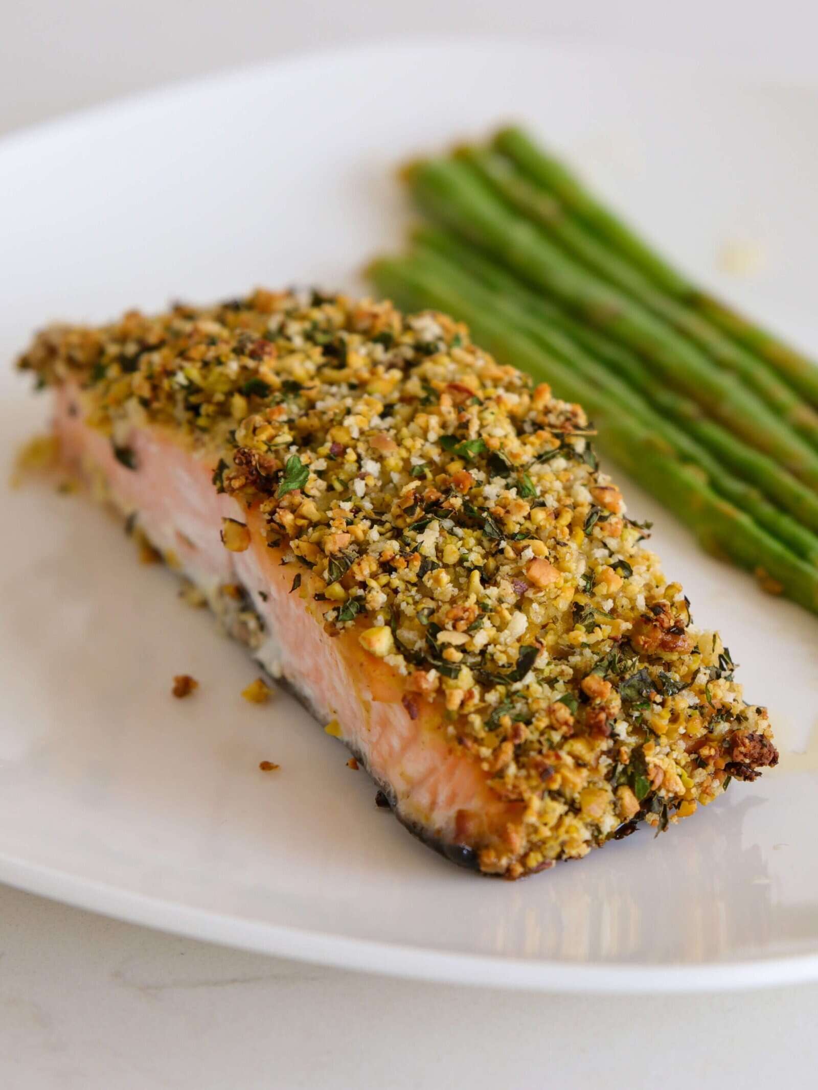 A piece of salmon with a golden herb and pistachio crust sits on a white plate next to several spears of cooked asparagus.